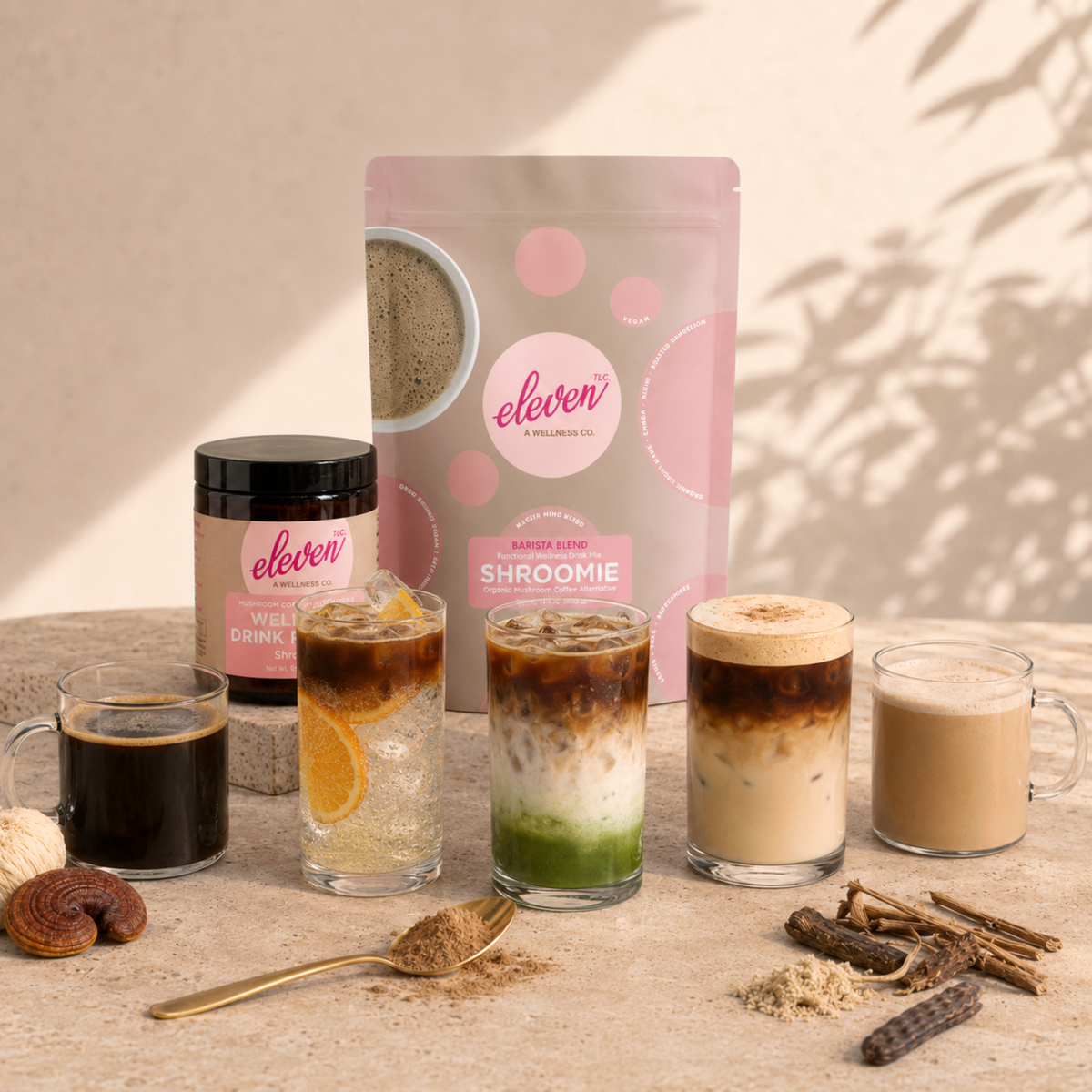 Eleven TLC Shroomie Barista Blend 500g pouch and 168g amber glass jar behind five Shroomie drinks: a black brew, an iced tonic with orange, an iced matcha layer drink, an iced latte and a hot latte, with a gold spoon of powder and ingredients on travertine.