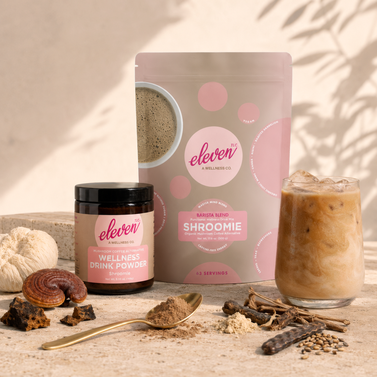Eleven TLC Shroomie Barista Blend 500g pouch and 168g amber glass jar beside an iced latte, with lion’s mane, reishi, chaga, carob and a gold spoon of powder on a travertine